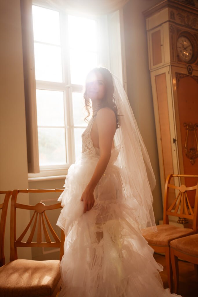 the bride is standing next to a big window, with sunlight flasing thrugh