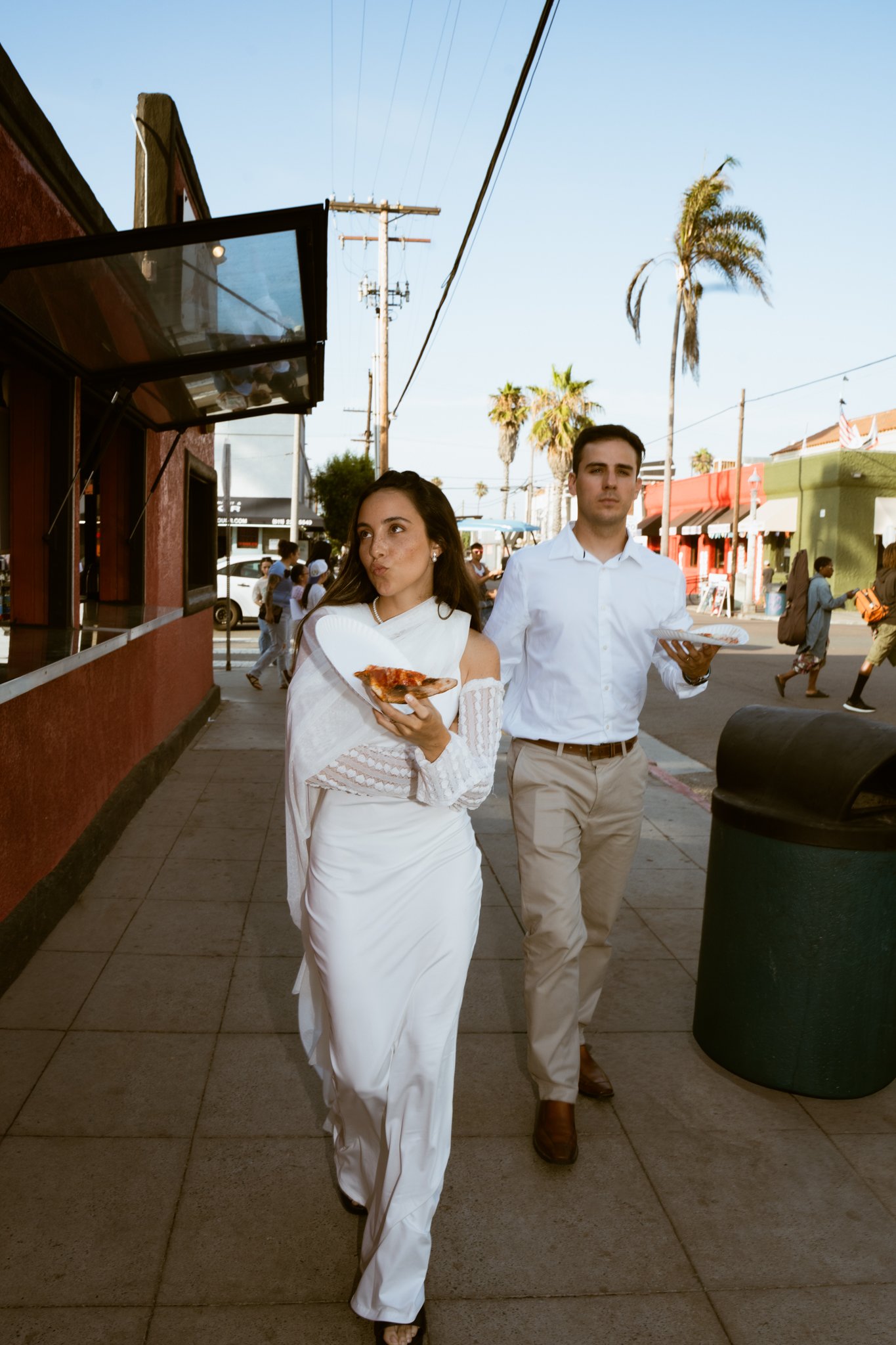 bride eating pizza in weddingdress, groom walking behind