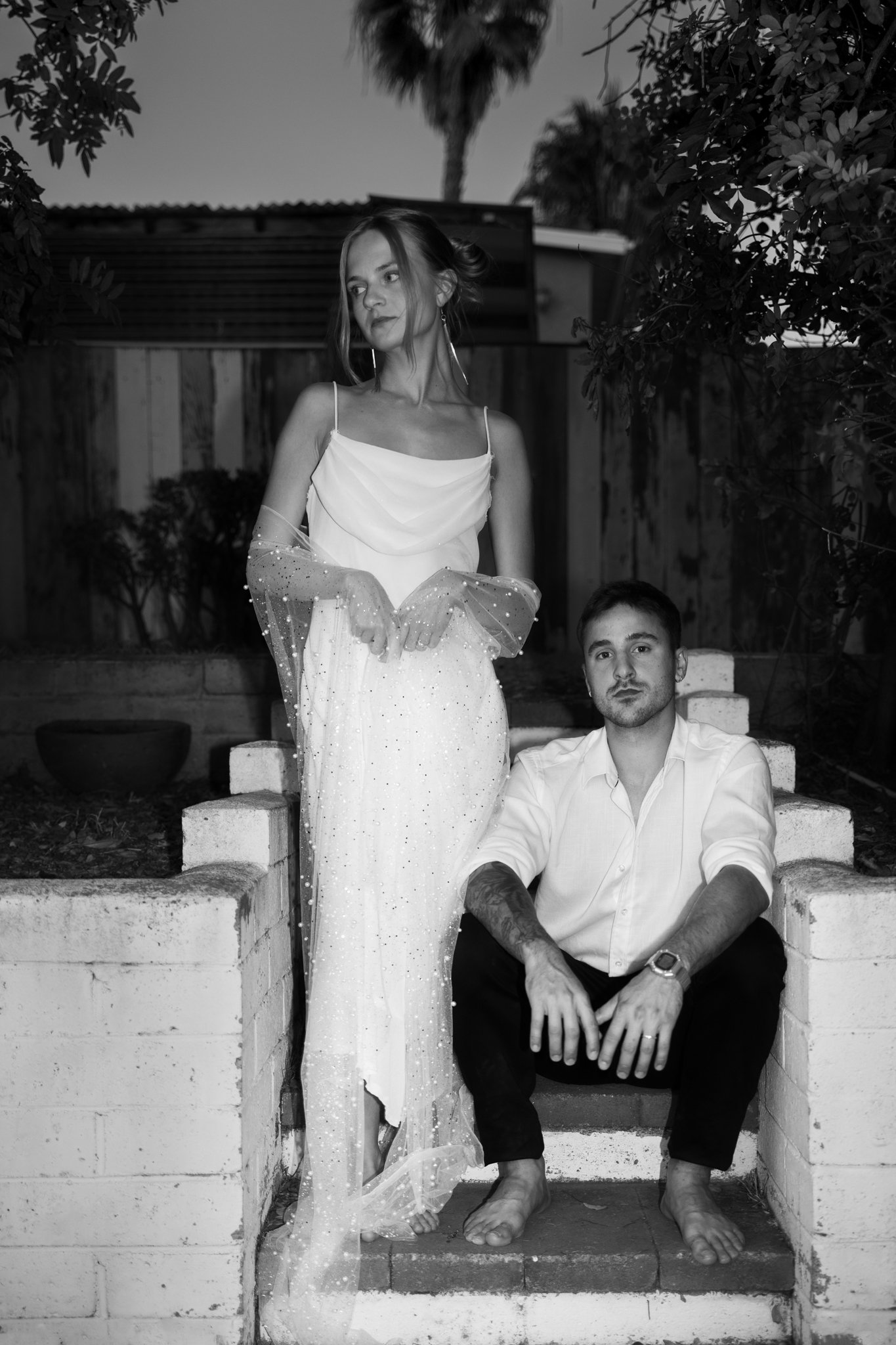 Woman in Weddingdress standing on stairs, Man sitting beside her and looking straight to camera.